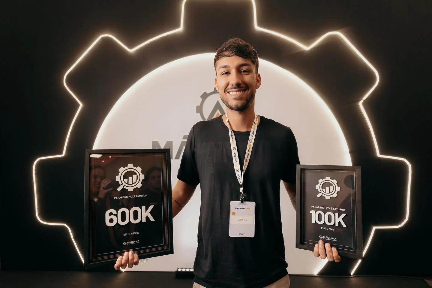André Tini sorrindo num evento, segurando duas placas de faturamento, uma de 600 mil e outra de 100 mil